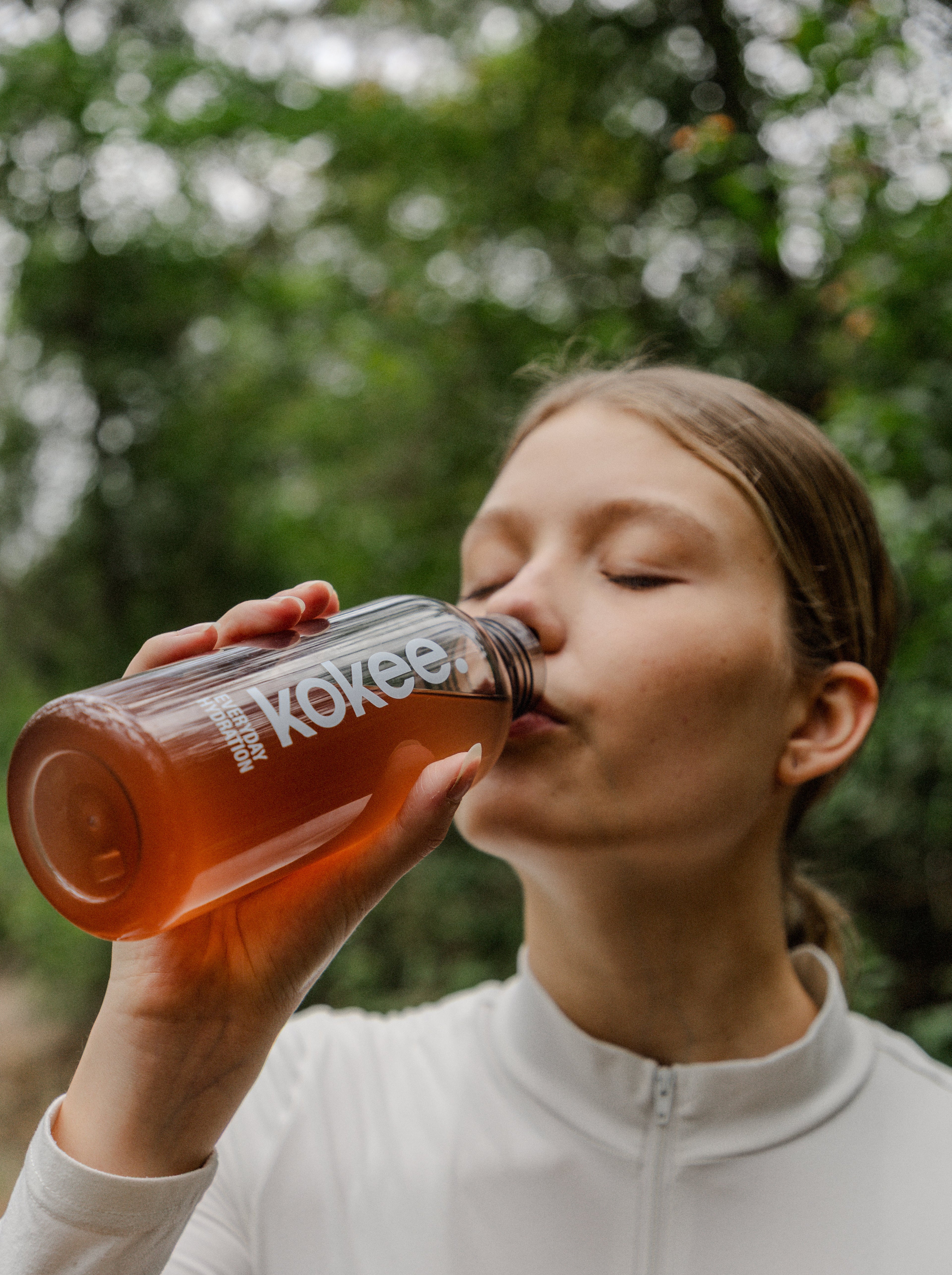Person drinking from a Kokee bottle outdoors with greenery in the background